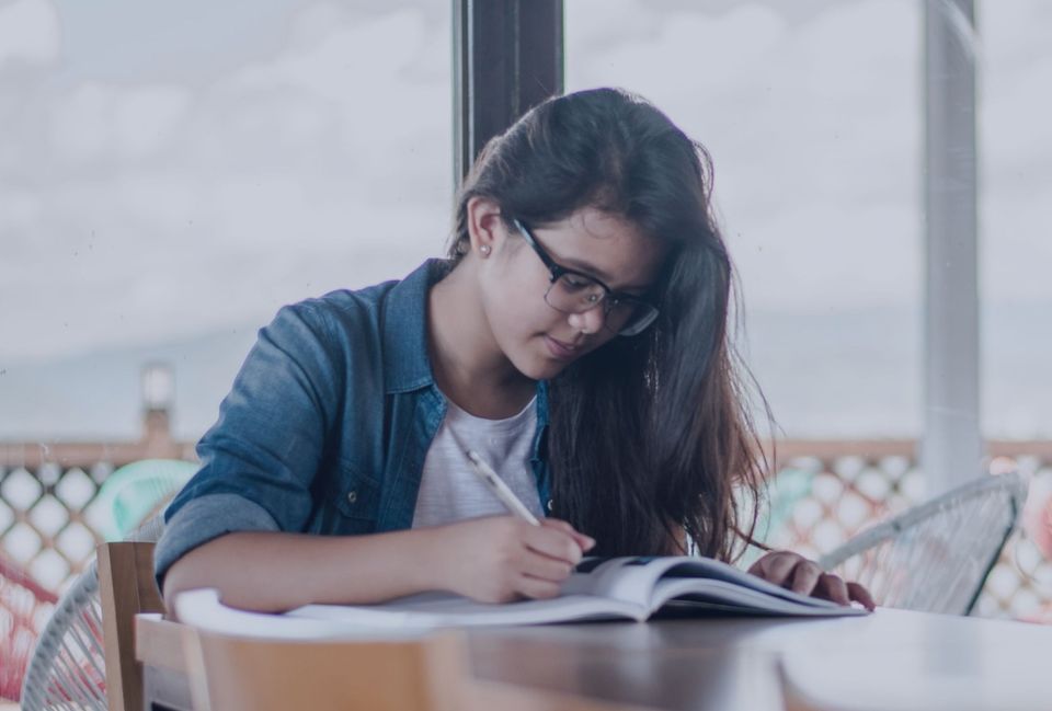 Person with long dark hair wearing glasses and a denim shirt writing in an open notebook, with grey skies in the background