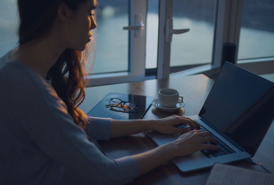 Person with long dark hair typing on a laptop near an open window, with rays of sunset on their face